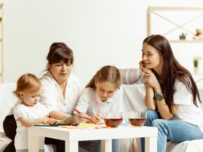 Two little girls their attractive young mother and their charming grandmother sitting on sofa and spending time together at home. Generation of women. International Women's Day. Happy Mother's Day.